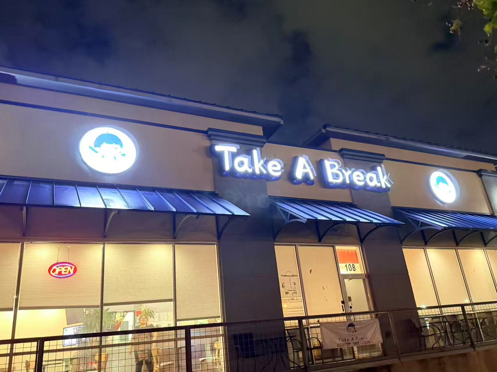 Night view of "Take A Break" café, with bright neon sign, glowing "OPEN" light, and warm interior visible through large windows.