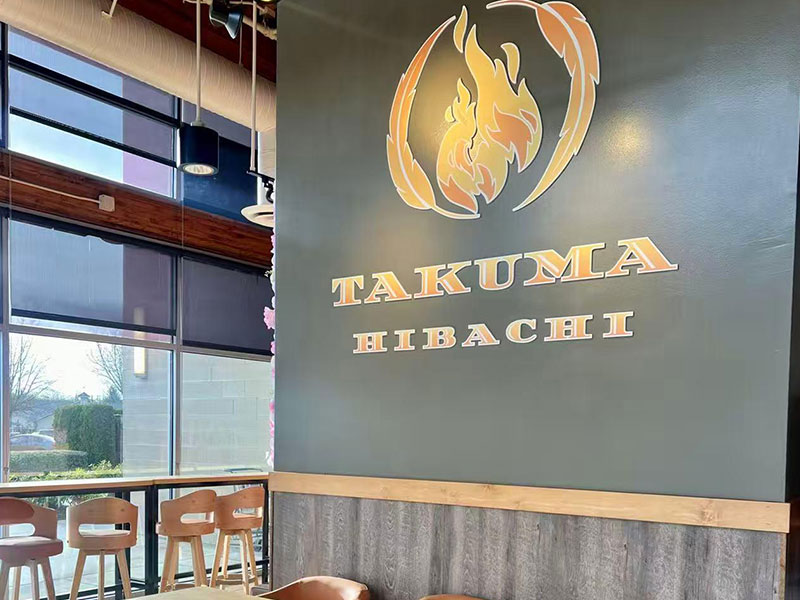 Interior wall featuring golden Takuma Hibachi flame logo beside large glass windows and wooden dining stools at Takuma Hibachi , a Japanese Restaurant in Hillsboro