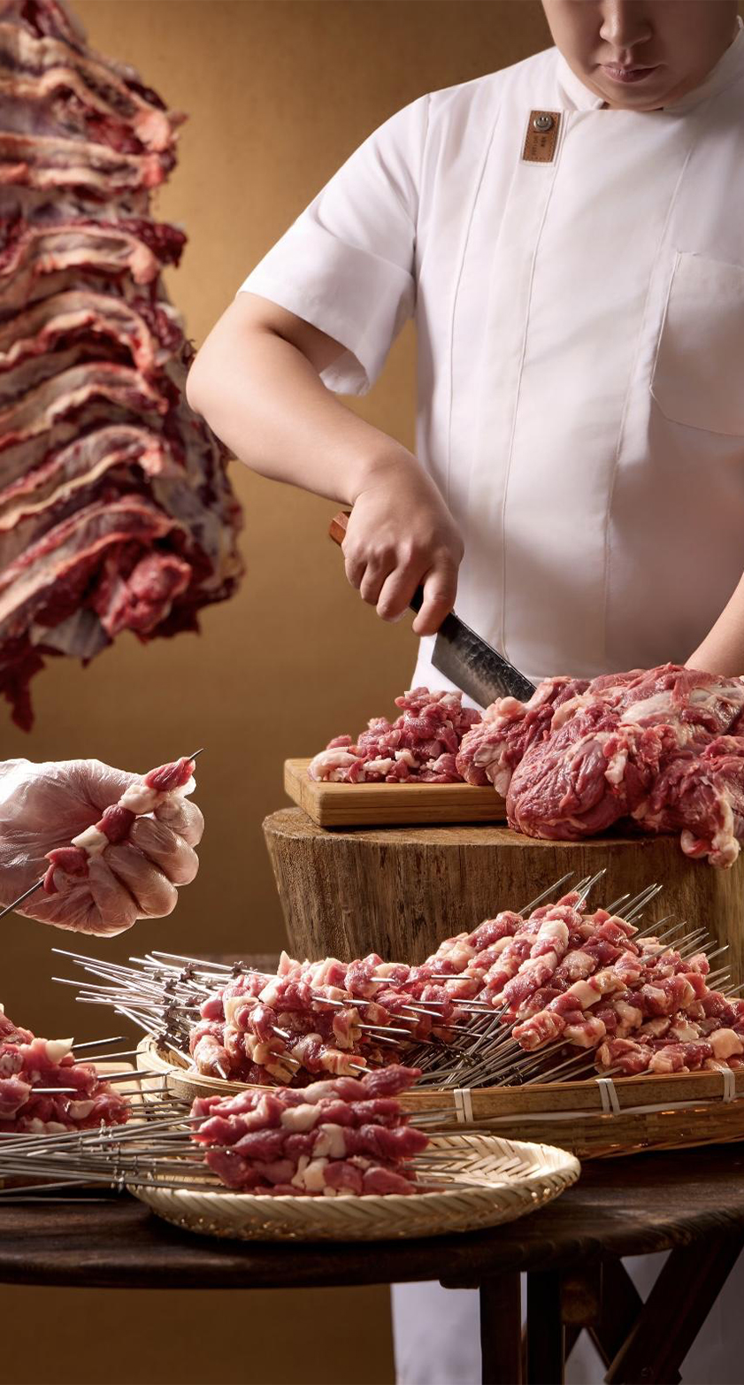 A chef cutting fresh meat on a wooden stump surrounded by prepared raw meat skewers