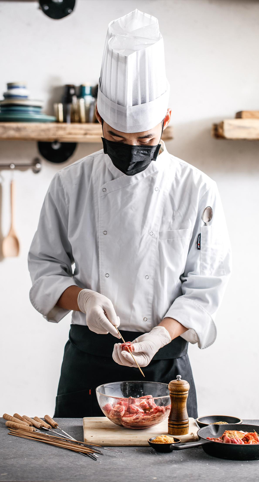 Professional chef in a mask and gloves carefully skewering raw meat pieces onto sticks