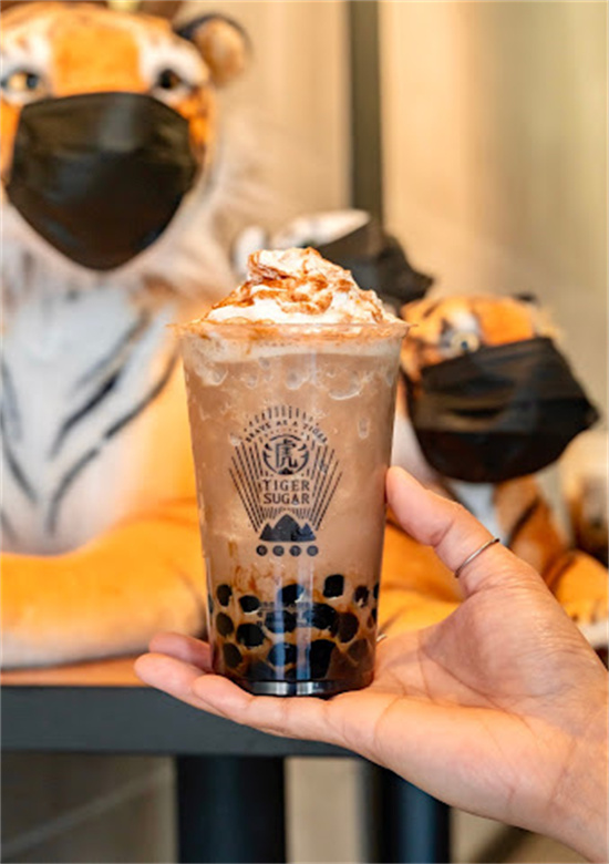 Hand holding whipped cream-topped boba drink in front of masked tiger plush toys at Tiger Sugar, a Bubble tea store in Philadelphia