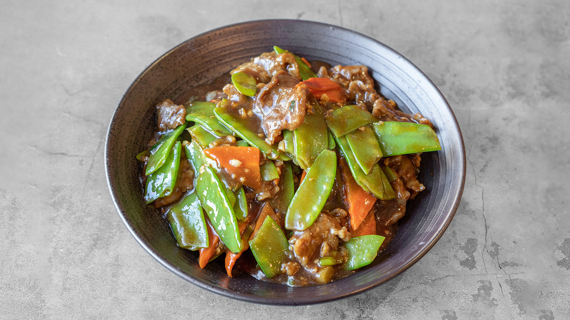 Stir-fried beef with snow peas and carrot slices coated in thick brown savory sauce in dark speckled bowl at Mandarin Chinese & Japanese Cuisine , a Chinese Restaurant in Santa Monica
