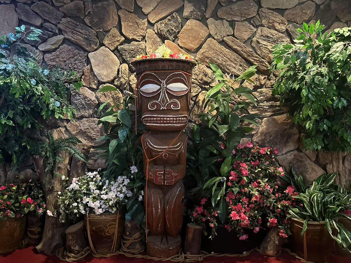 A decorative Tiki statue surrounded by potted plants, flowers, and greenery against a stone wall at Tong's Tiki Hut，a Asian Fusion Restaurant in Villa Park