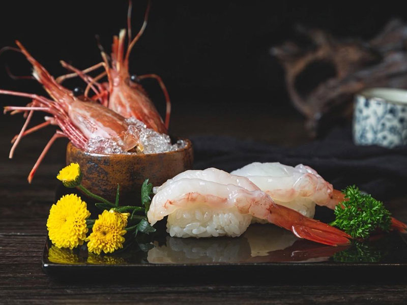 Two pieces of giant shrimp nigiri sushi on dark reflective plate with raw shrimp in ceramic bowl and yellow floral garnish at Toromi Sushi , a Sushi Restaurant in West Covina