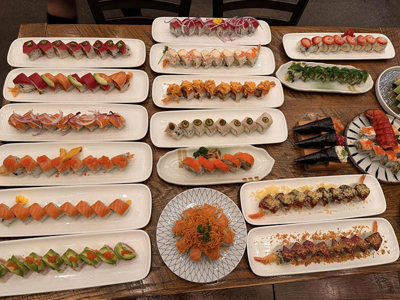 Large wooden table spread with dozens of assorted sushi rolls, hand rolls and sashimi platters in white rectangular plates at Toromi Sushi , a Sushi Restaurant in West Covina