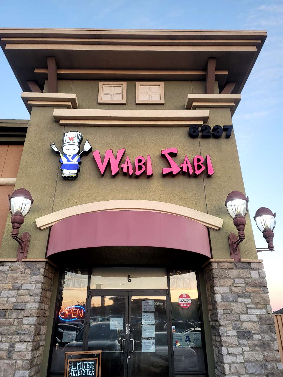 Wabi-Sabi-Japanese-Restaurant-Jurupa-Valley The storefront of "Wabi Sabi" features a pink logo, a cartoon chef mascot, and an "OPEN" neon sign above the glass doors.
