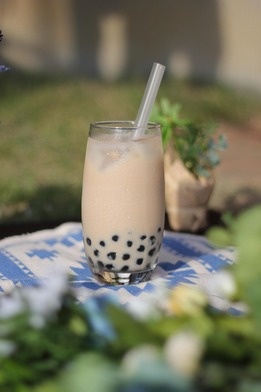 Glass of bubble tea with tapioca pearls and a straw, placed on a patterned cloth outdoors with greenery in soft focus background.