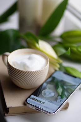 Beige ceramic cup of frothy coffee on a book beside a smartphone displaying a plant photo, with green leaves in soft background.