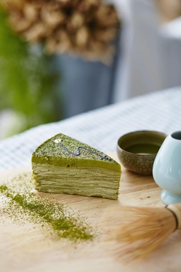 Slice of layered green matcha crepe cake dusted with matcha powder, served on a wooden board with a small bowl of tea and a blue pitcher.