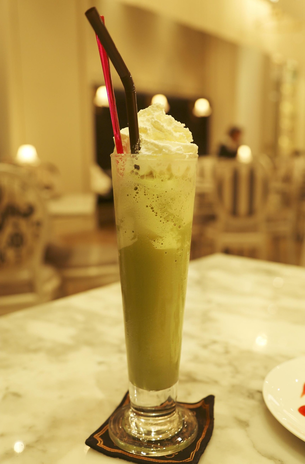 Tall glass of iced matcha latte topped with whipped cream and two straws, placed on a marble table in an elegant cafe setting.