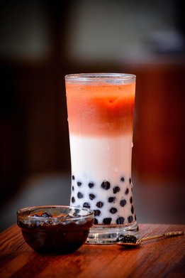 Tall glass of layered Thai iced tea with tapioca pearls, accompanied by a small bowl of boba and a spoon on a wooden table.