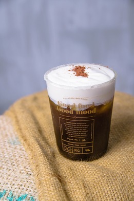 Clear plastic cup of iced coffee topped with thick cream foam and cocoa powder, sitting on a woven burlap surface.