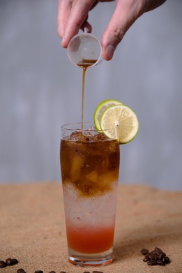 Hand pouring espresso into a tall glass of layered iced drink with lime slices on the rim, coffee beans scattered nearby.