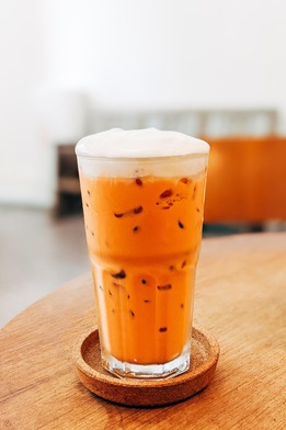 Tall glass of Thai iced tea with a frothy top layer, served on a round wooden coaster on a light wooden table.