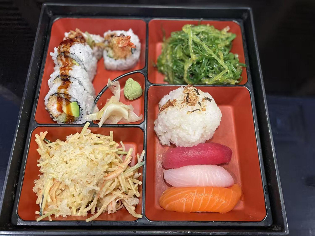A Japanese bento box divided into compartments, containing sushi rolls, seaweed salad, rice ball, and slices of raw fish at Yume Hibachi Steak and Sushi，a Japanese Restaurant in Unionville