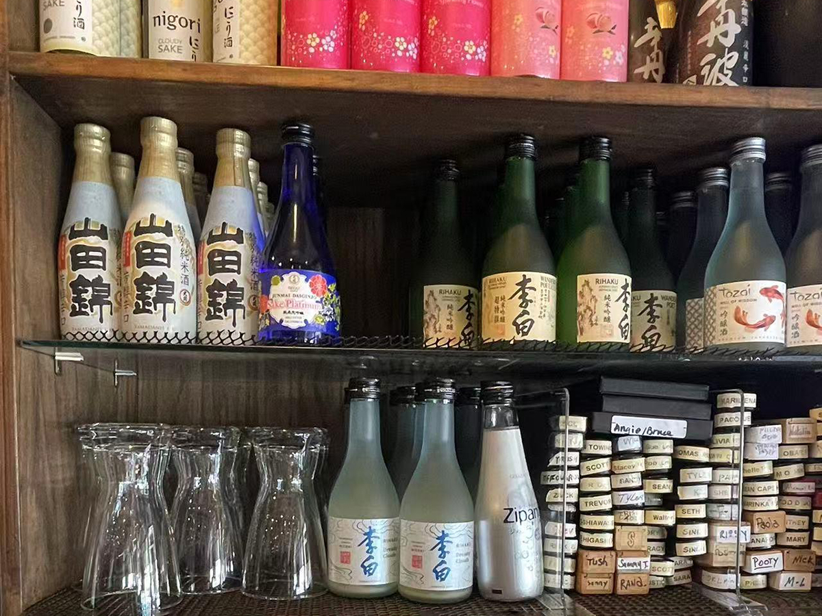 A shelf displaying various bottles of sake, with some sake glasses placed in front of the bottles at Yume Hibachi Steak and Sushi，a Japanese Restaurant in Unionville