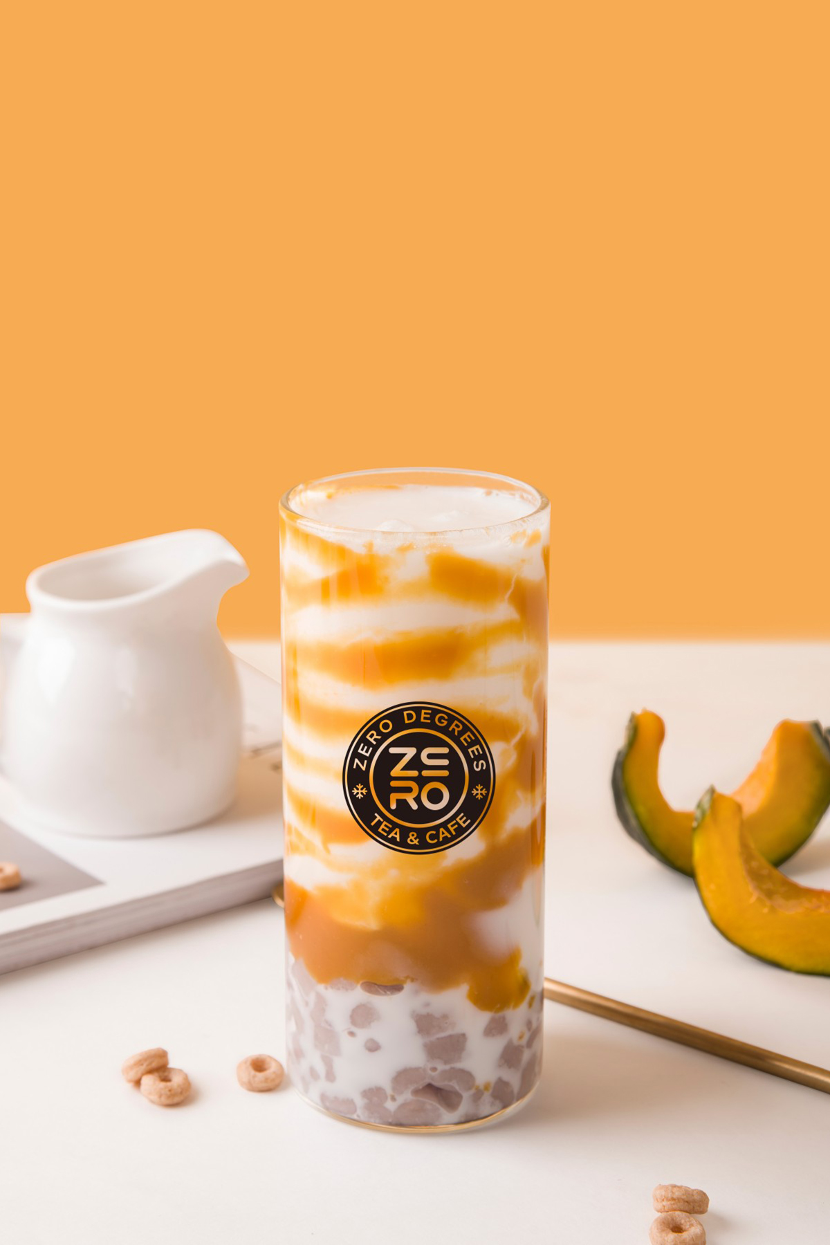 Zero-Degrees-Asian-inspired-Drink & Snack-Shop-Pumpkin-Taro-Ball-Milk-Tea-Bloomington A glass of pumpkin taro ball milk tea with pumpkin slices and cereal rings around, against an orange background at Zero Degrees,a Asian-inspired Drink & Snack Shop in Bloomington