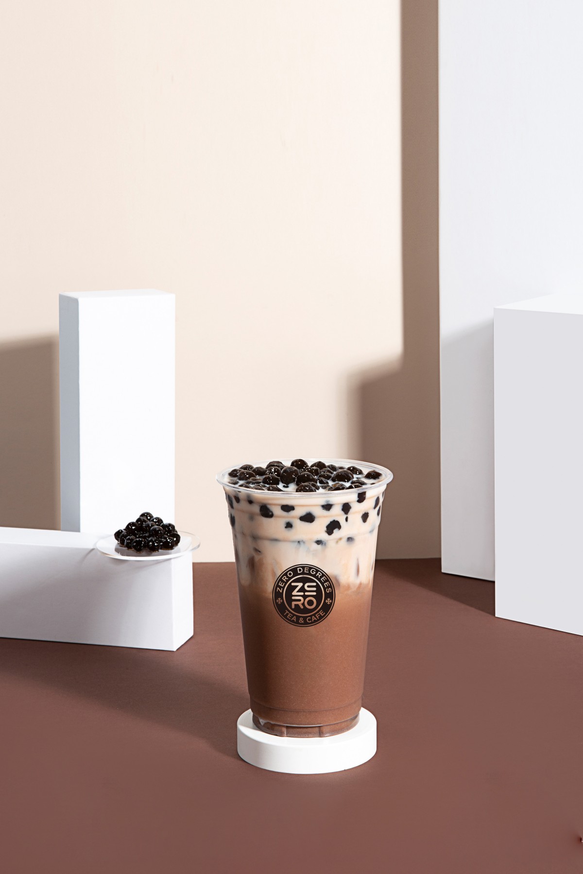 Zero-Degrees-Asian-inspired-Drink & Snack-Shop-Chocolate-Boba-Milk-Tea-Bloomington A cup of chocolate boba milk tea with boba on a small plate, placed among white geometric shapes on a brown surface at Zero Degrees,a Asian-inspired Drink & Snack Shop in Bloomington