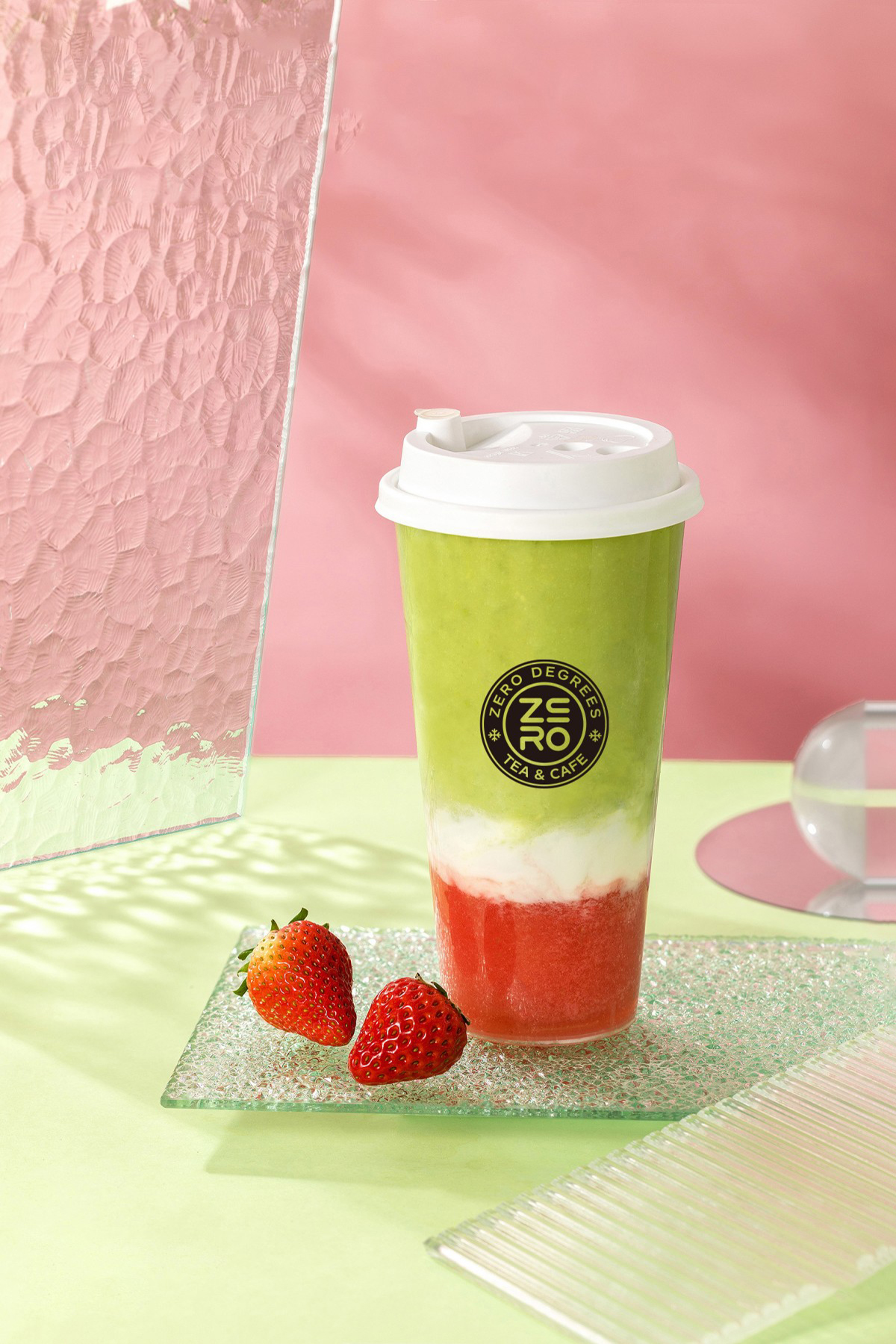 Zero-Degrees-Asian-inspired-Drink & Snack-Shop-Strawberry-Matcha-Layered-Milk-Cap-Tea-Bloomington A cup of strawberry matcha layered milk cap tea with two strawberries beside it, set against a pink and green background at Zero Degrees,a Asian-inspired Drink & Snack Shop in Bloomington