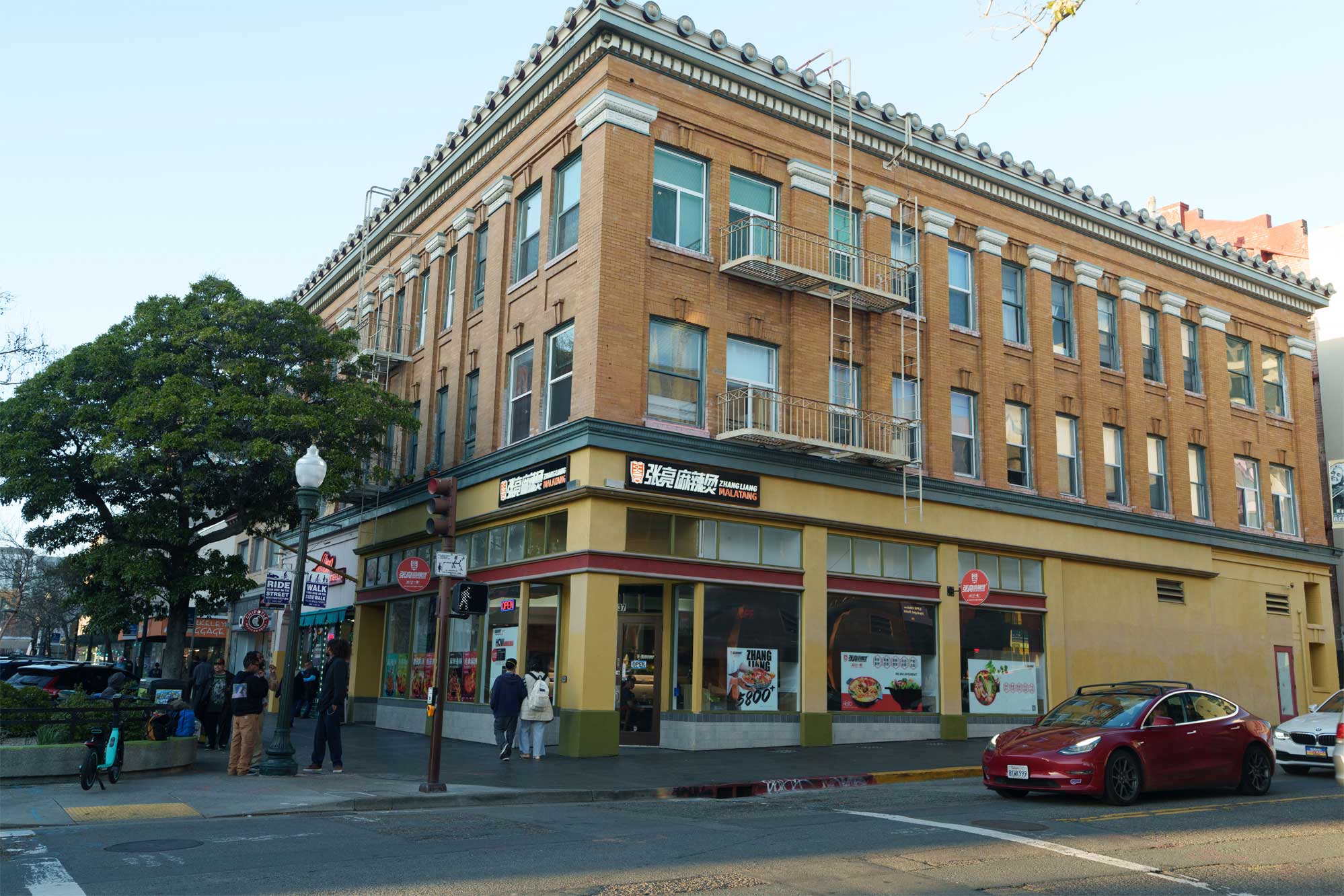 Street‑level view of Zhang Liang Malatang restaurant occupying ground floor of vintage multi‑story brick corner building, pedestrians on sidewalk, traffic light, red Tesla car on city street, large tree and street signs visible.